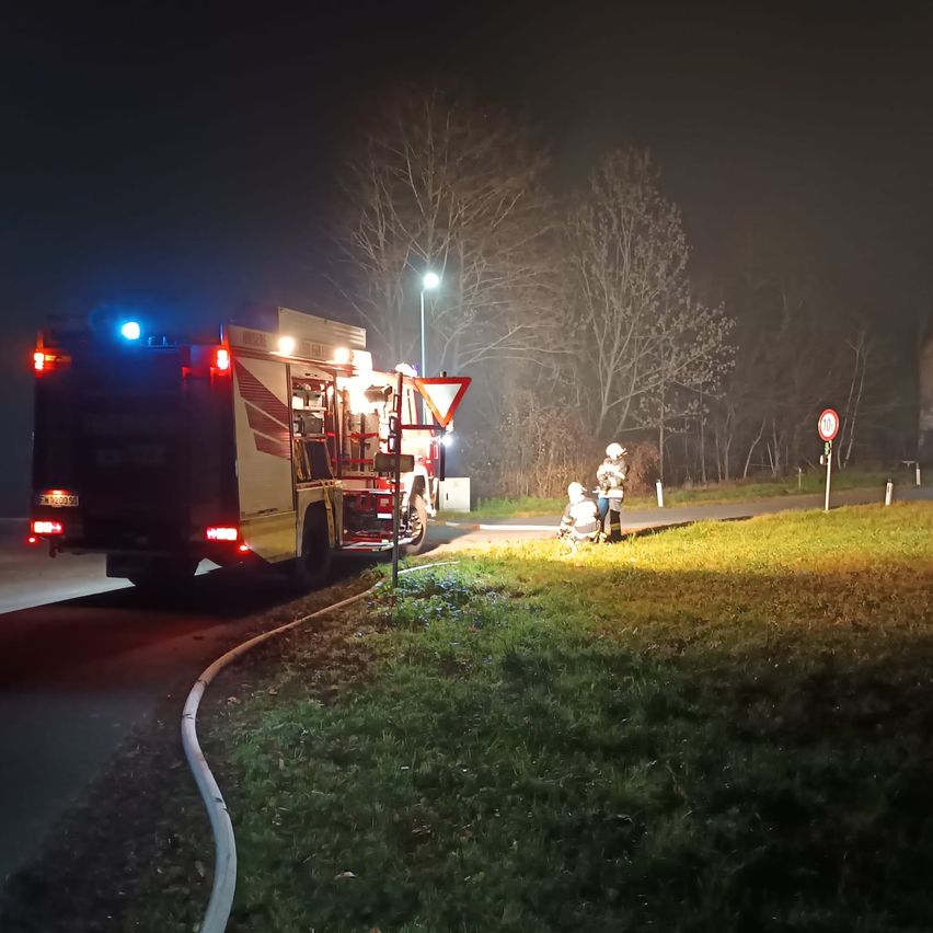 A fire truck with flashing lights is parked on the side of the road at night. Two people are standing on the grass, one in a white helmet.