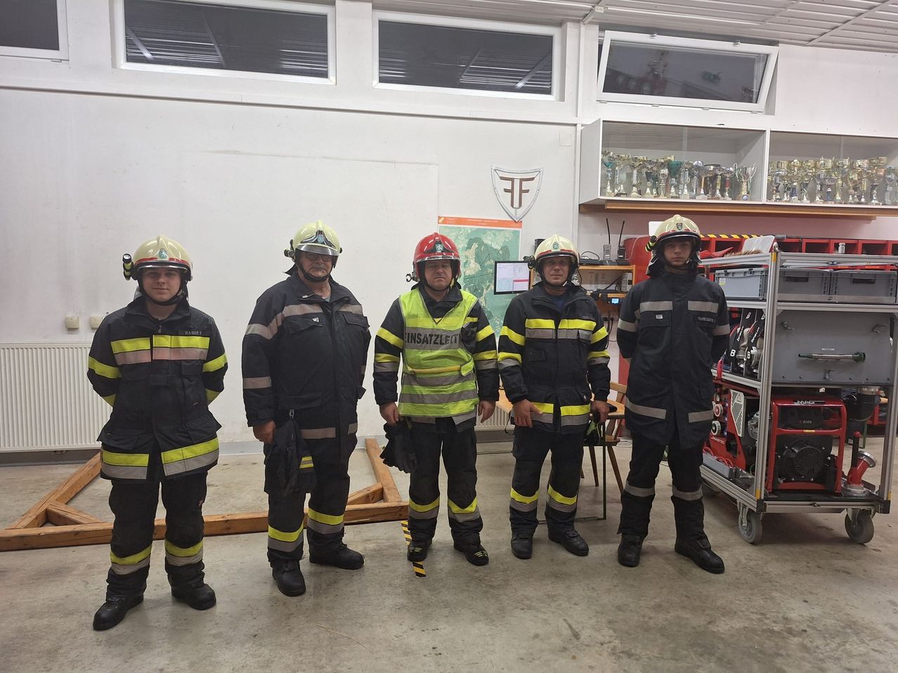 Five firefighters stand in a room with white walls and windows. They are wearing helmets, uniforms, and reflective strips. In the background, trophies are displayed on a shelf.