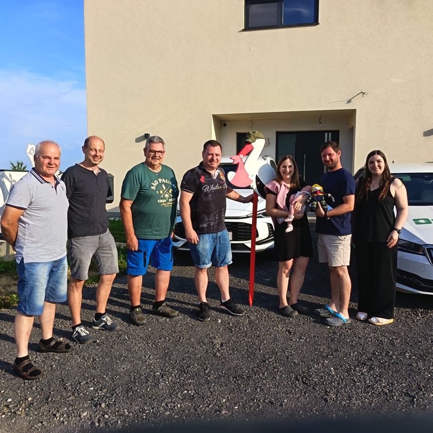 Eine Gruppe von Menschen steht vor einem Gebäude mit einem geparkten weißen Auto. Sie lächeln und posieren für ein Foto. In der Mitte der Gruppe befindet sich ein Storch-Spielzeug.