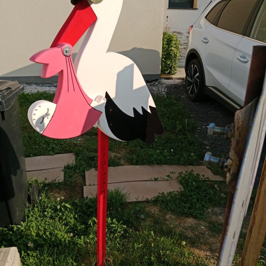 Ein rosa und weißer Storchbriefkasten steht im Garten neben einem weißen Auto und einem Mülleimer. Der Storch hält ein Baby im Schnabel.
