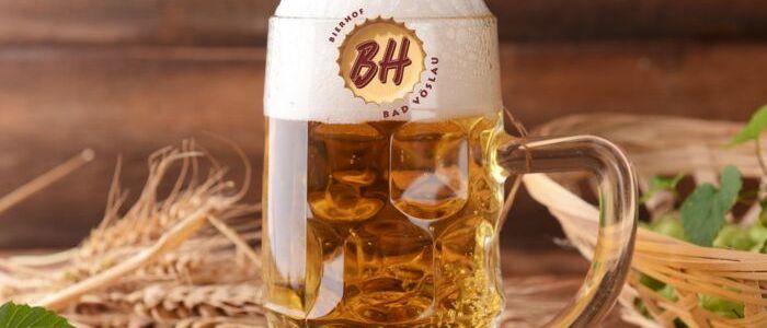 Ein Glas Bier mit Schaum, mit dem Etikett Bierhof Bad Vöslau, umgeben von Hopfen und Weizen auf einem Holztisch.