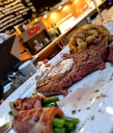 Ein weißer Teller mit Steak, Kartoffelpüree, grünen Bohnen und einem Weinglas. Der Restauranttresen ist im Hintergrund verschwommen.