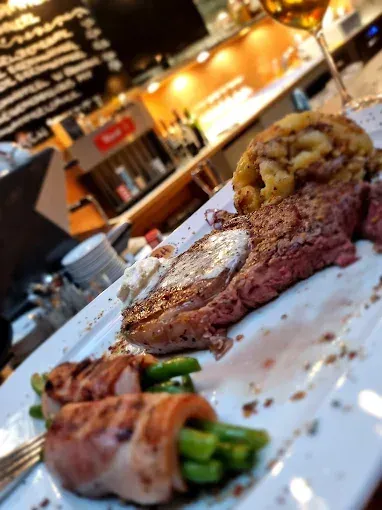 Ein weißer Teller mit Steak, Kartoffelpüree, grünen Bohnen und einem Weinglas. Der Restauranttresen ist im Hintergrund verschwommen.