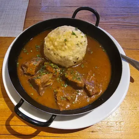 Eine Pfanne mit Rindergulasch mit einem großen Knödel steht auf einem weißen Teller auf einem Holztisch.