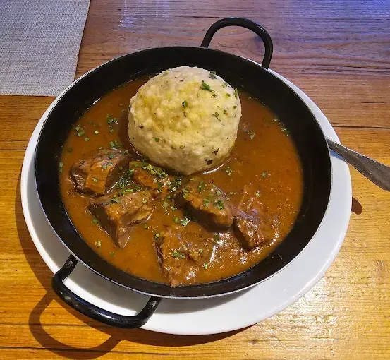 Eine Pfanne mit Rindergulasch mit einem großen Knödel steht auf einem weißen Teller auf einem Holztisch.