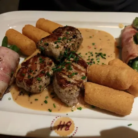 Ein weißer Teller enthält ein Gericht mit gegrilltem Fleisch, grünen Bohnen und gebratenen Frühlingsrollen. Das Gericht hat eine cremige Sauce und Kräuter.