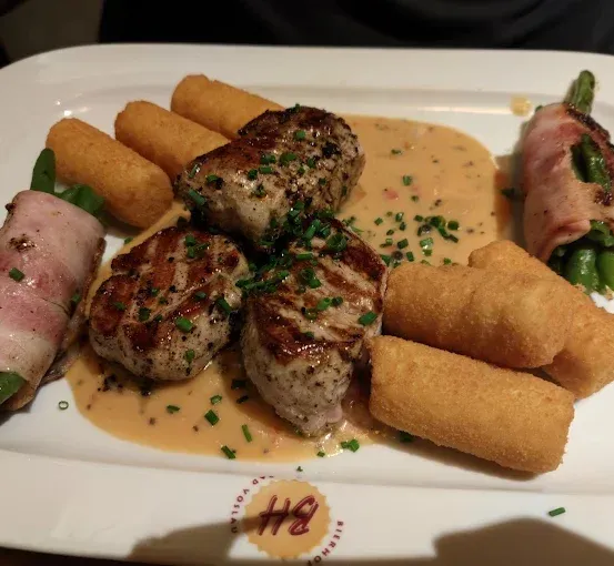 Ein weißer Teller enthält ein Gericht mit gegrilltem Fleisch, grünen Bohnen und gebratenen Frühlingsrollen. Das Gericht hat eine cremige Sauce und Kräuter.