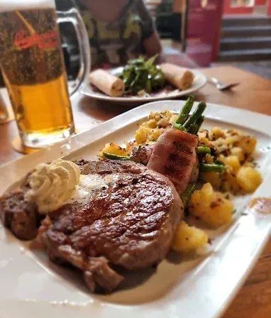 Ein Steak mit cremiger Sauce, grünen Bohnen und Kartoffeln steht auf einem Teller. Ein Bierkrug und ein weiterer Teller mit Essen sind auf dem Tisch.