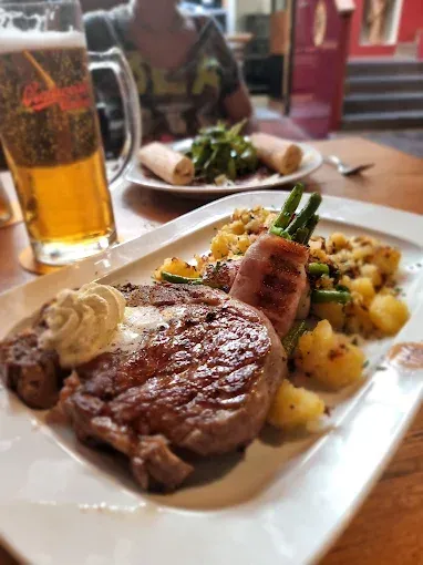 Ein Steak mit cremiger Sauce, grünen Bohnen und Kartoffeln steht auf einem Teller. Ein Bierkrug und ein weiterer Teller mit Essen sind auf dem Tisch.