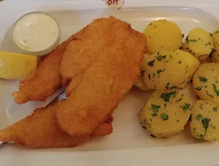 Ein weißer Teller mit gebratenen Fischstücken, gekochten Kartoffeln, einer Zitronenscheibe und einem Klecks saure Sahne auf einem Holztisch.