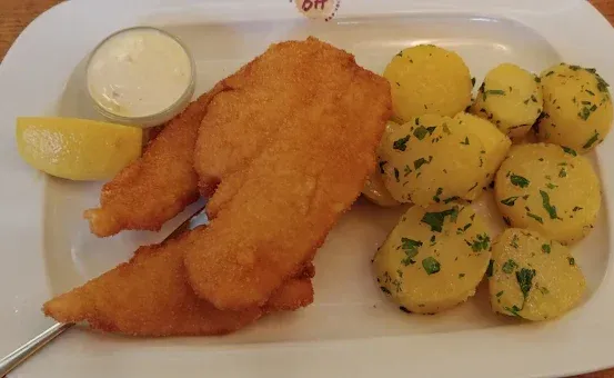 Ein weißer Teller mit gebratenen Fischstücken, gekochten Kartoffeln, einer Zitronenscheibe und einem Klecks saure Sahne auf einem Holztisch.