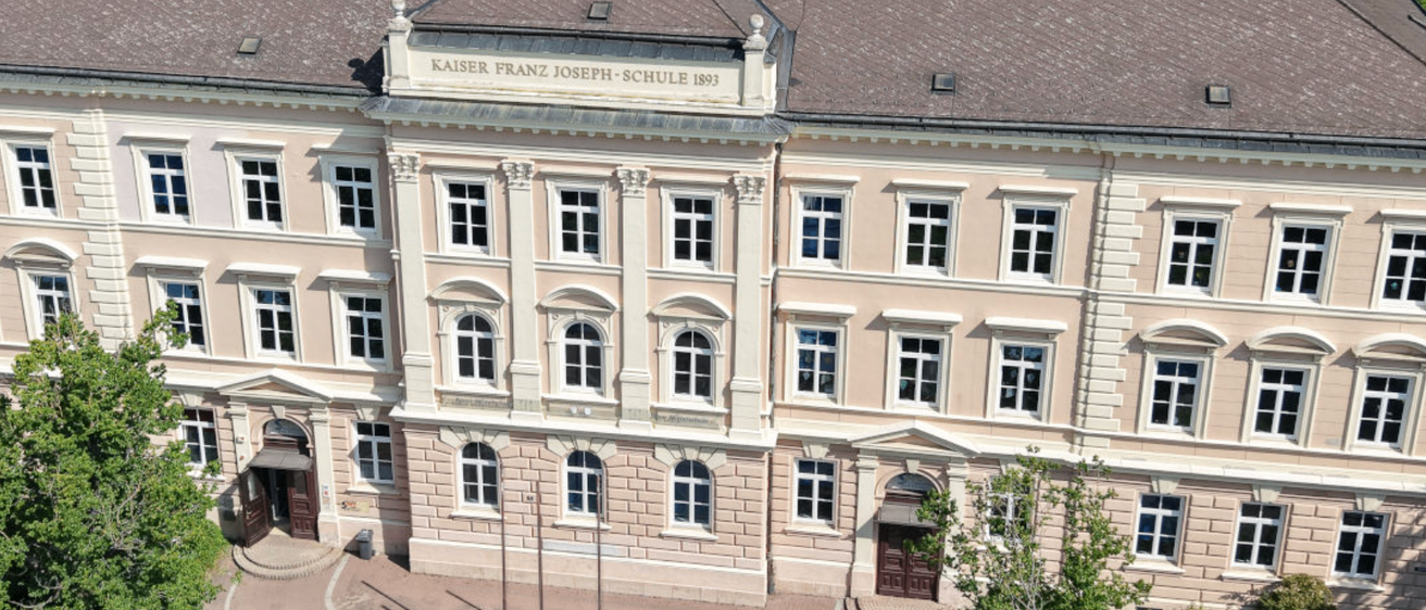 Ein Luftbild der Kaiser Franz Joseph-Schule, ein Gebäude mit einem braunen Dach und vielen Fenstern.