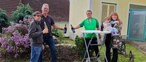 Eine Gruppe von Menschen posiert für ein Foto im Garten, mit einem Haus im Hintergrund. Sie halten Flaschen und ein Baby.