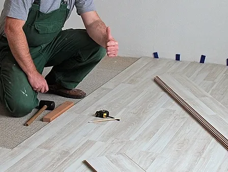 Ein Mann in grüner Arbeitskleidung kniet auf dem Boden und installiert wahrscheinlich einen Laminatboden.