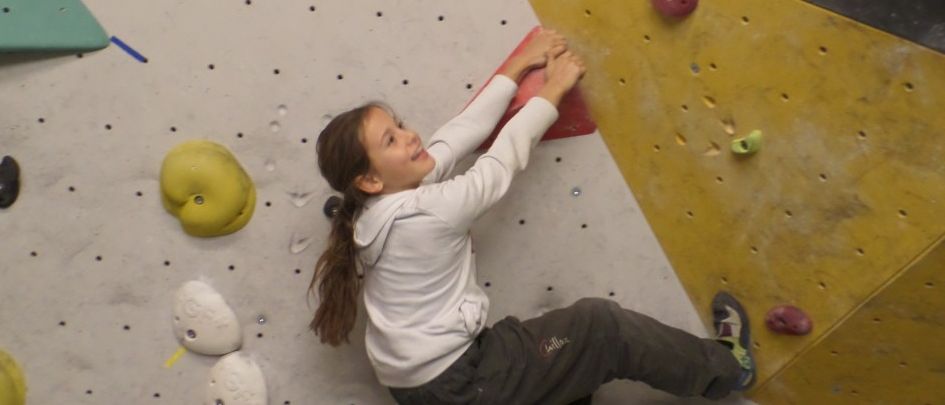 Ein Mädchen in einer weißen Kapuzenjacke klettert eine Kletterwand mit verschiedenen Griffen und Haltepunkten. Die Wand ist mehrfarbig mit gelben, roten und grauen Abschnitten.