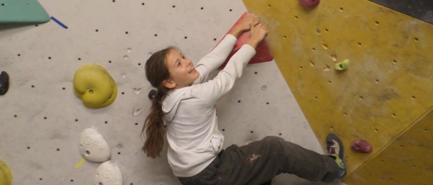 Ein Mädchen in einer weißen Kapuzenjacke klettert eine Kletterwand mit verschiedenen Griffen und Haltepunkten. Die Wand ist mehrfarbig mit gelben, roten und grauen Abschnitten.