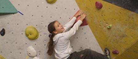 Ein junges Mädchen klettert an einer Indoor-Kletterwand. Sie trägt ein weißes Sweatshirt und graue Hose. Verschiedene farbige Klettergriffe sind an der Wand befestigt.