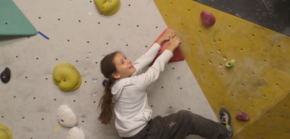 Ein junges Mädchen klettert an einer Indoor-Kletterwand. Sie trägt ein weißes Sweatshirt und graue Hose. Verschiedene farbige Klettergriffe sind an der Wand befestigt.