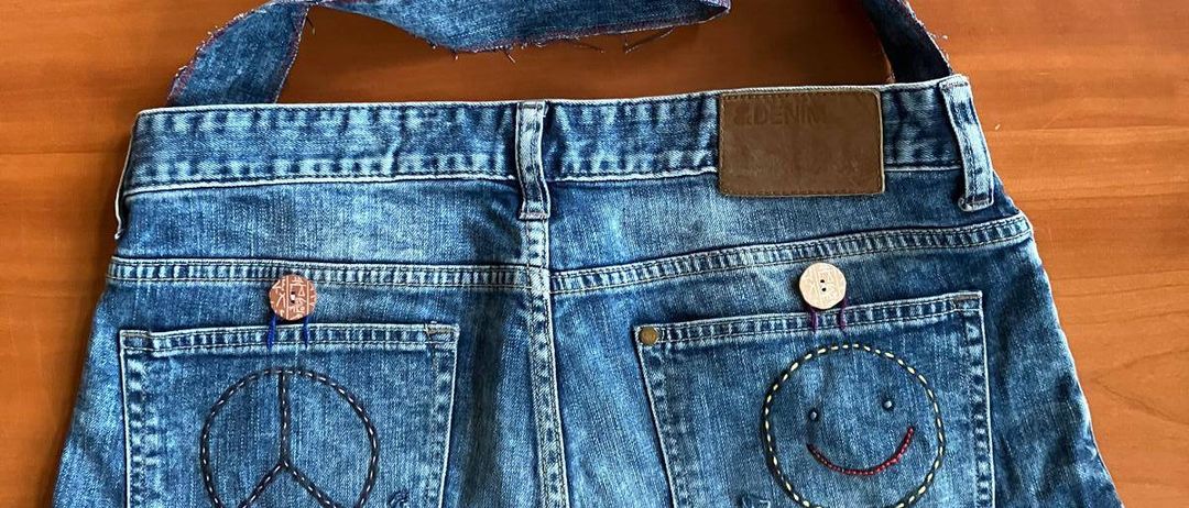 Eine Jeanstasche mit Friedenszeichen und lächelndem Gesicht auf den Gesäßtaschen, einem braunen Etikett und einem Henkel liegt auf einer Holzoberfläche.
