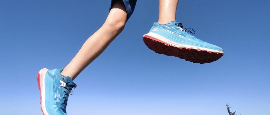 Eine Person in blauer Shorts und blauen Turnschuhen springt über einen Grashügel in die Luft, mit blauem Himmel im Hintergrund.