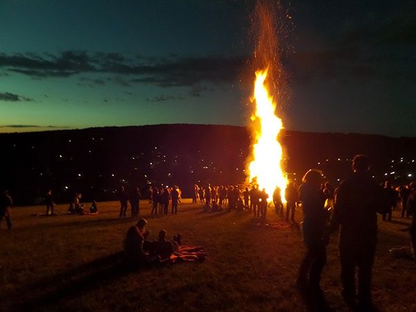 Ein großes Lagerfeuer bei Nacht mit Menschen, die sich darum versammelt haben und auf dem Boden sitzen. Das Feuer brennt hell und wirft warmes Licht auf die Menschen und die Umgebung. In der Ferne gibt es Hügel und eine Stadtlandschaft mit Lichtern.
