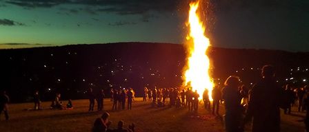 Ein großes Lagerfeuer bei Nacht mit Menschen, die sich darum versammelt haben und auf dem Boden sitzen. Das Feuer brennt hell und wirft warmes Licht auf die Menschen und die Umgebung. In der Ferne gibt es Hügel und eine Stadtlandschaft mit Lichtern.