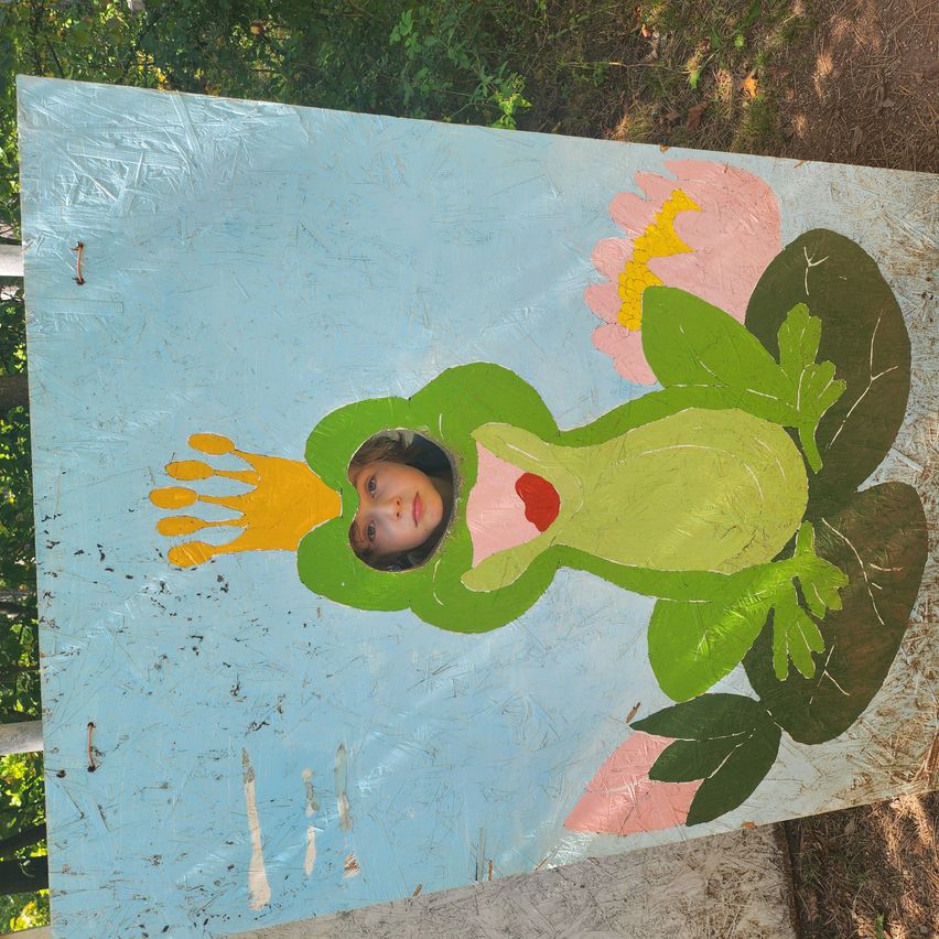 Ein handbemaltes Holzschild zeigt einen grünen Frosch, aus dessen Auge ein Kind herausschaut, umgeben von rosa Blumen und grünen Blättern, auf einer Holzoberfläche.