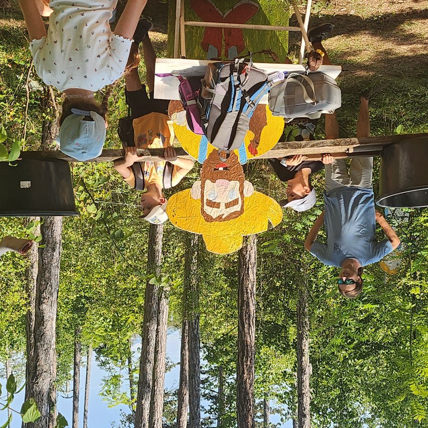 Eine Gruppe von Kindern und Erwachsenen steht kopfüber in einem Wald, einer hält eine große gelbe Figur.