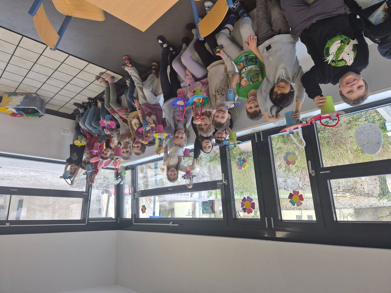 A classroom full of children hanging upside down from the ceiling, holding colorful toys, with large windows behind them.