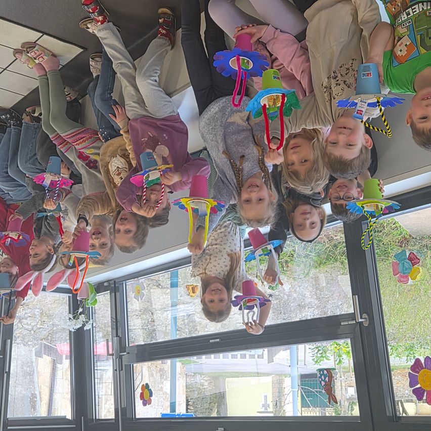 Children standing upside down, holding colorful crafts above their heads. Some wear glasses. Windows reflect buildings outside.