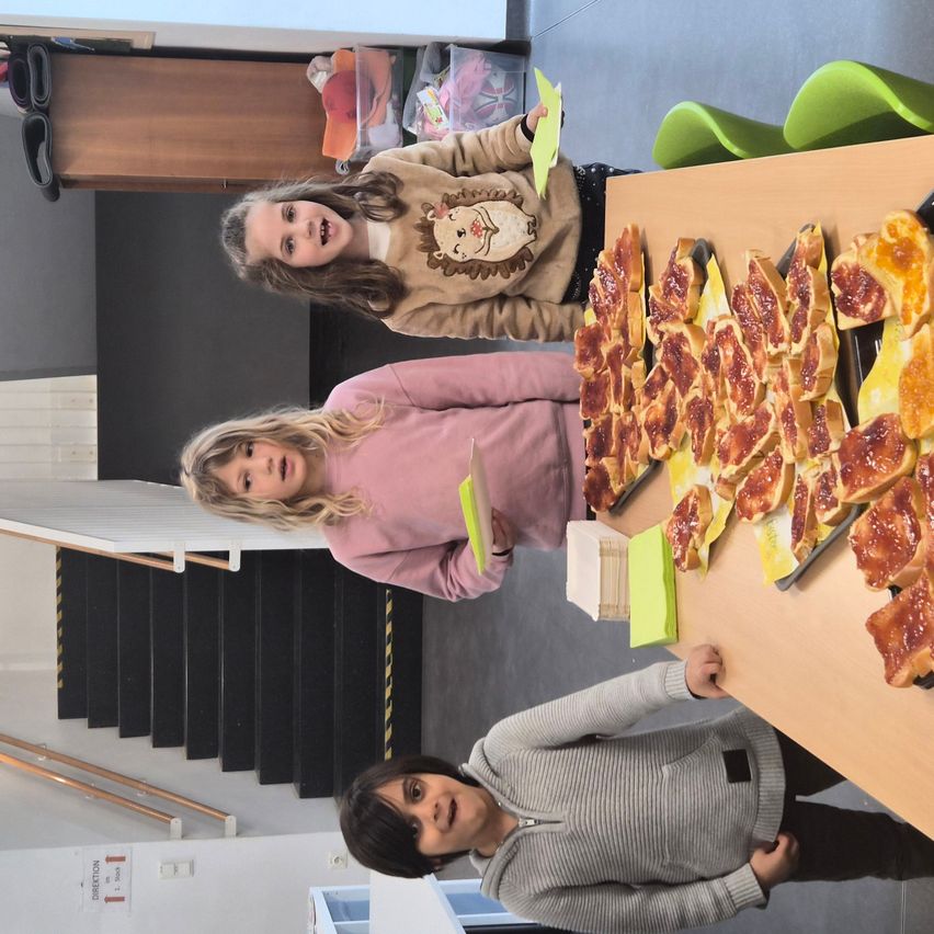 Three children stand by a table with food. Two of them hold paper, and the third reaches for a slice of bread. A staircase is in the background.