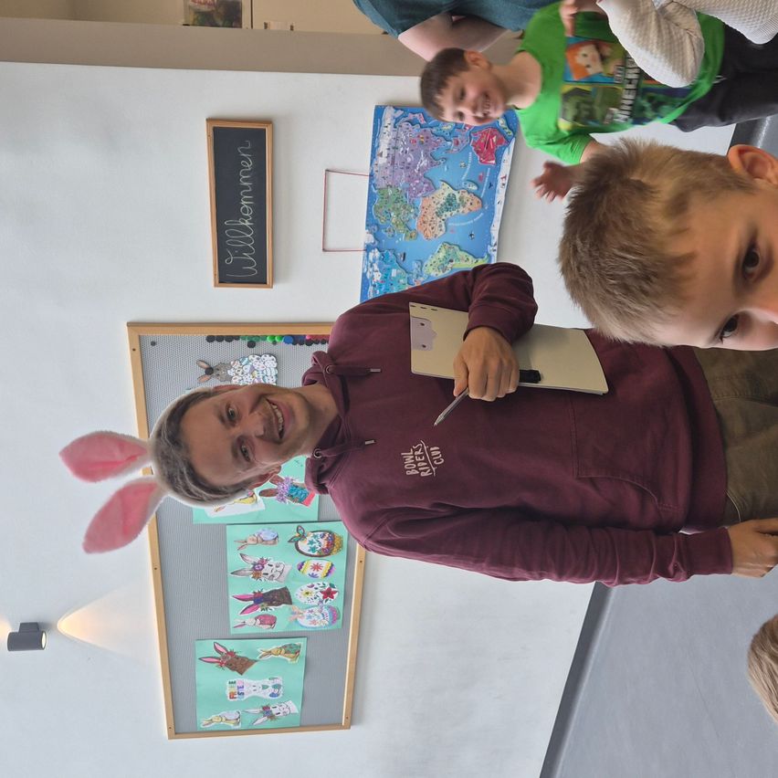 A man in bunny ears writes on a clipboard while smiling. Behind him, children look on. The room has white walls, a bulletin board, and a blackboard.