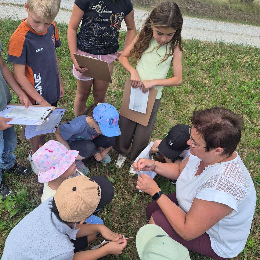 A group of children and an adult are gathered on a grassy field, possibly for a nature study. The children are holding clipboards and papers, and the adult is holding a magnifying glass.