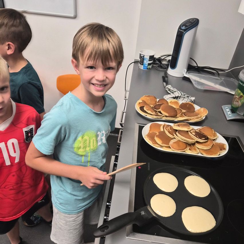 A young boy is standing in front of a stove, holding a wooden spatula, and smiling. There are two plates of pancakes on the stove and a cup on the counter.