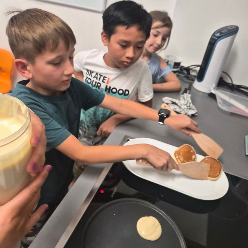 Three children are in a kitchen, one boy is flipping pancakes on a pan, another boy is watching, and a girl is sitting at the table.