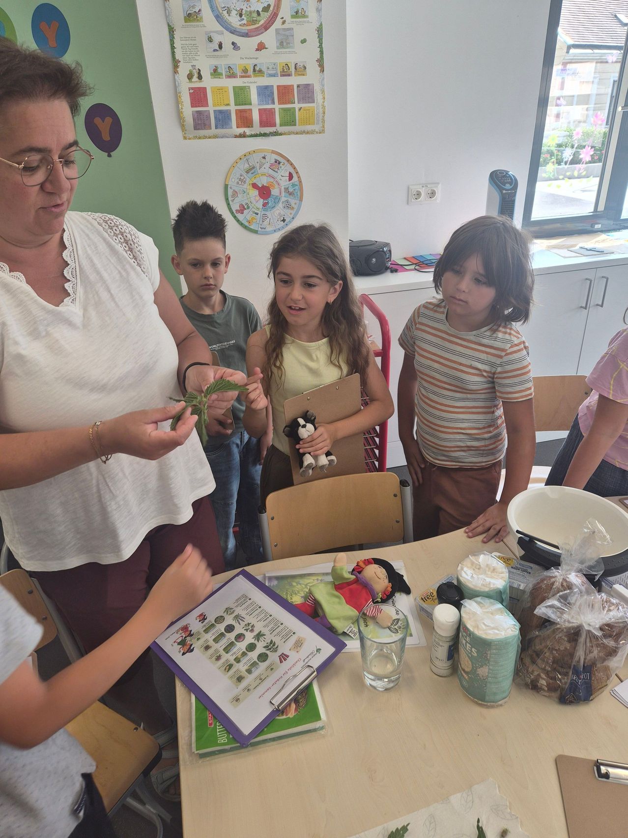 A woman holds a plant while explaining to a group of children, who are looking at a table with various items and a book.