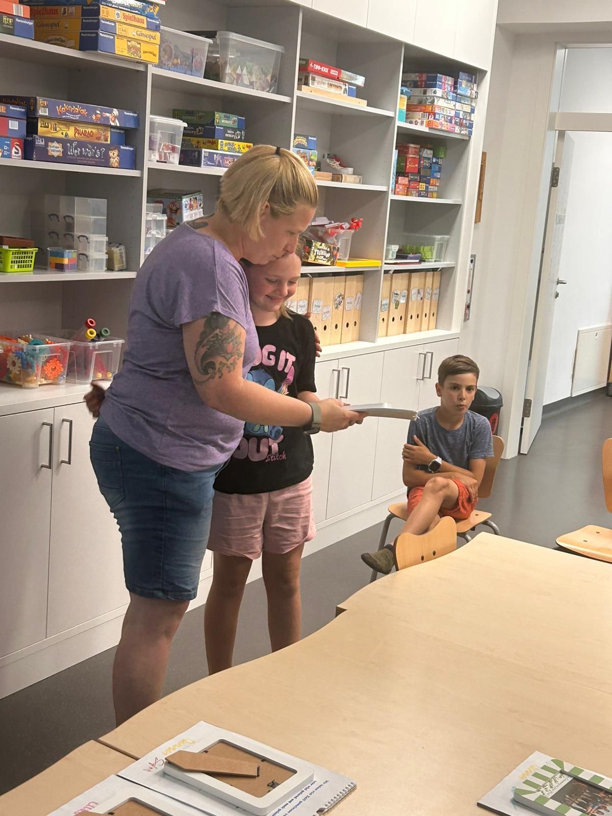 A woman hands a paper to a girl in a classroom while a boy watches. Behind them are shelves with toys and storage boxes.