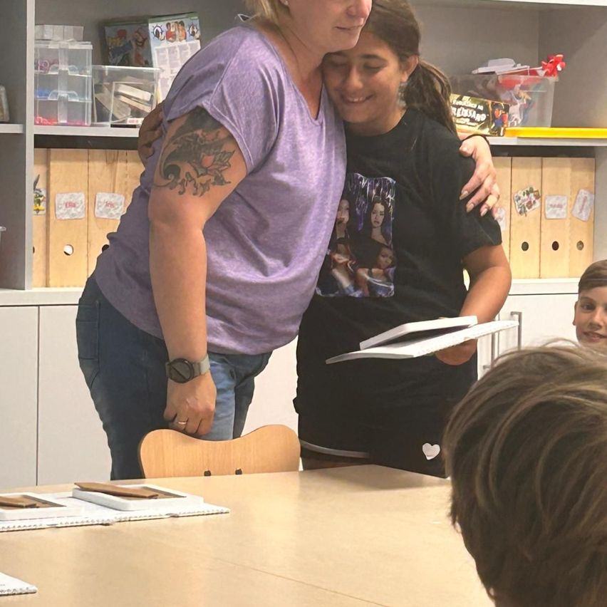 Two women hug in a classroom with a shelf and books in the background. One wears a purple shirt with a tattoo, the other a black shirt with a white heart.