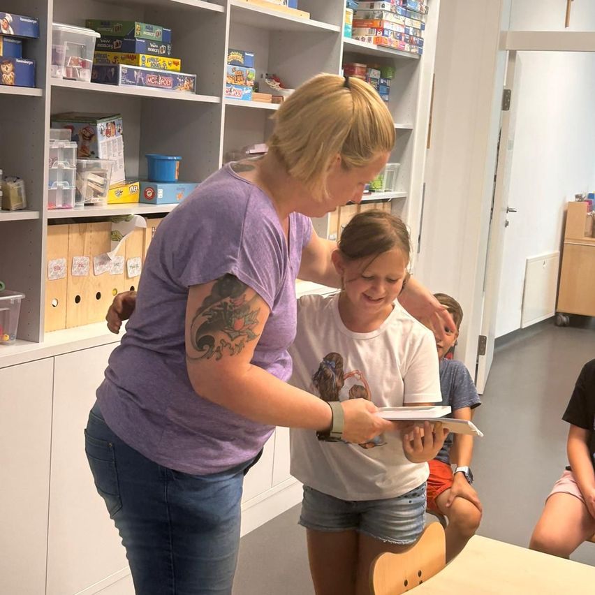 A woman with tattoos holds a young girl, smiling and holding a book. They stand in front of shelves filled with toys. Behind them, a boy sits on the floor.