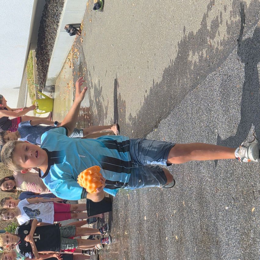 A young boy in a blue shirt holds a large orange sponge, standing in a driveway with several children around him. Shadows are cast on the ground.