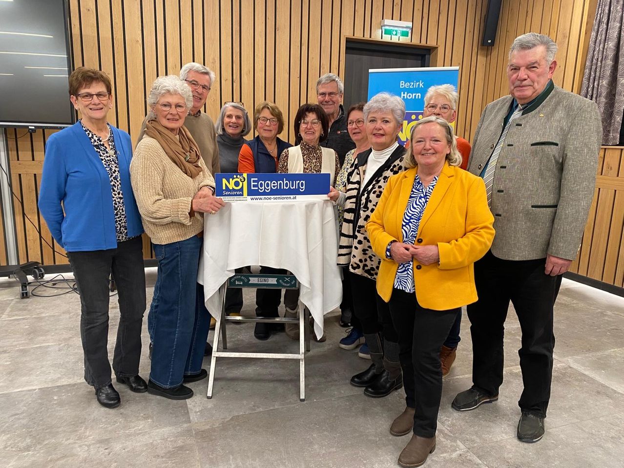 Eine Gruppe von Senioren steht hinter einem Tisch und hält ein blaues Schild mit der Aufschrift 'Eggenburg'. Sie lächeln und posieren für ein Foto. Hinter ihnen ist ein blaues Banner mit weißem Text zu sehen.