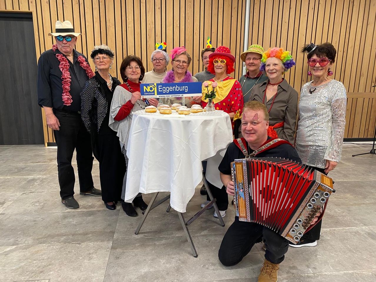 A group of elderly people dressed in various costumes and hats are posing for a photo, with a man playing an accordion and a table with pastries in the center.