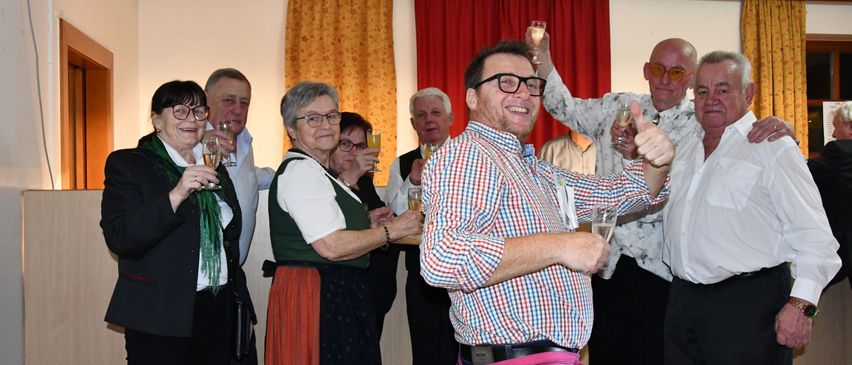 A group of adults, some wearing glasses, are standing together and holding glasses of wine. They appear to be celebrating. A man in the middle is wearing glasses and a plaid shirt, and he is smiling and giving a thumbs up.