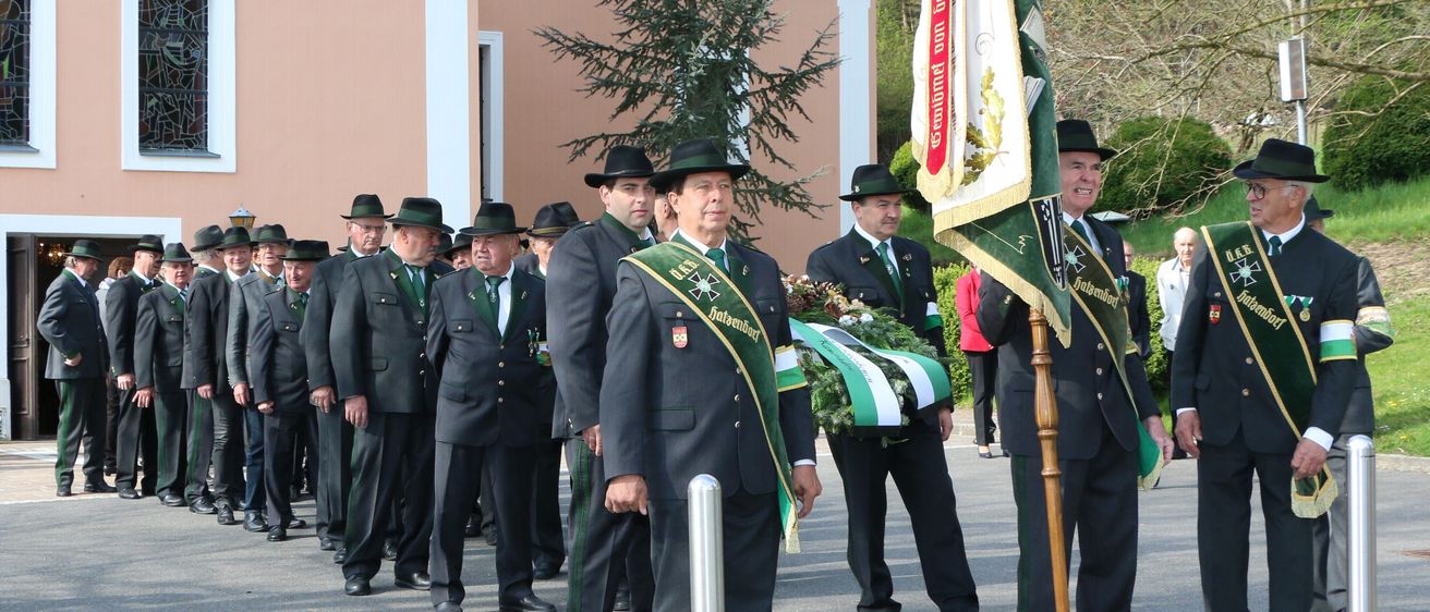 Eine Gruppe uniformierter Männer, möglicherweise Feuerwehrleute, steht draußen. Ein Mann vorne hält eine Kranz und scheint zu sprechen. Hinter ihm hält ein anderer Mann eine Fahne. Sie tragen alle Mützen und grüne Schärpen.