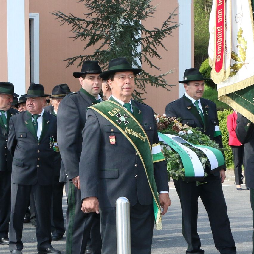 Eine Gruppe uniformierter Männer, möglicherweise Feuerwehrleute, steht draußen. Ein Mann vorne hält eine Kranz und scheint zu sprechen. Hinter ihm hält ein anderer Mann eine Fahne. Sie tragen alle Mützen und grüne Schärpen.
