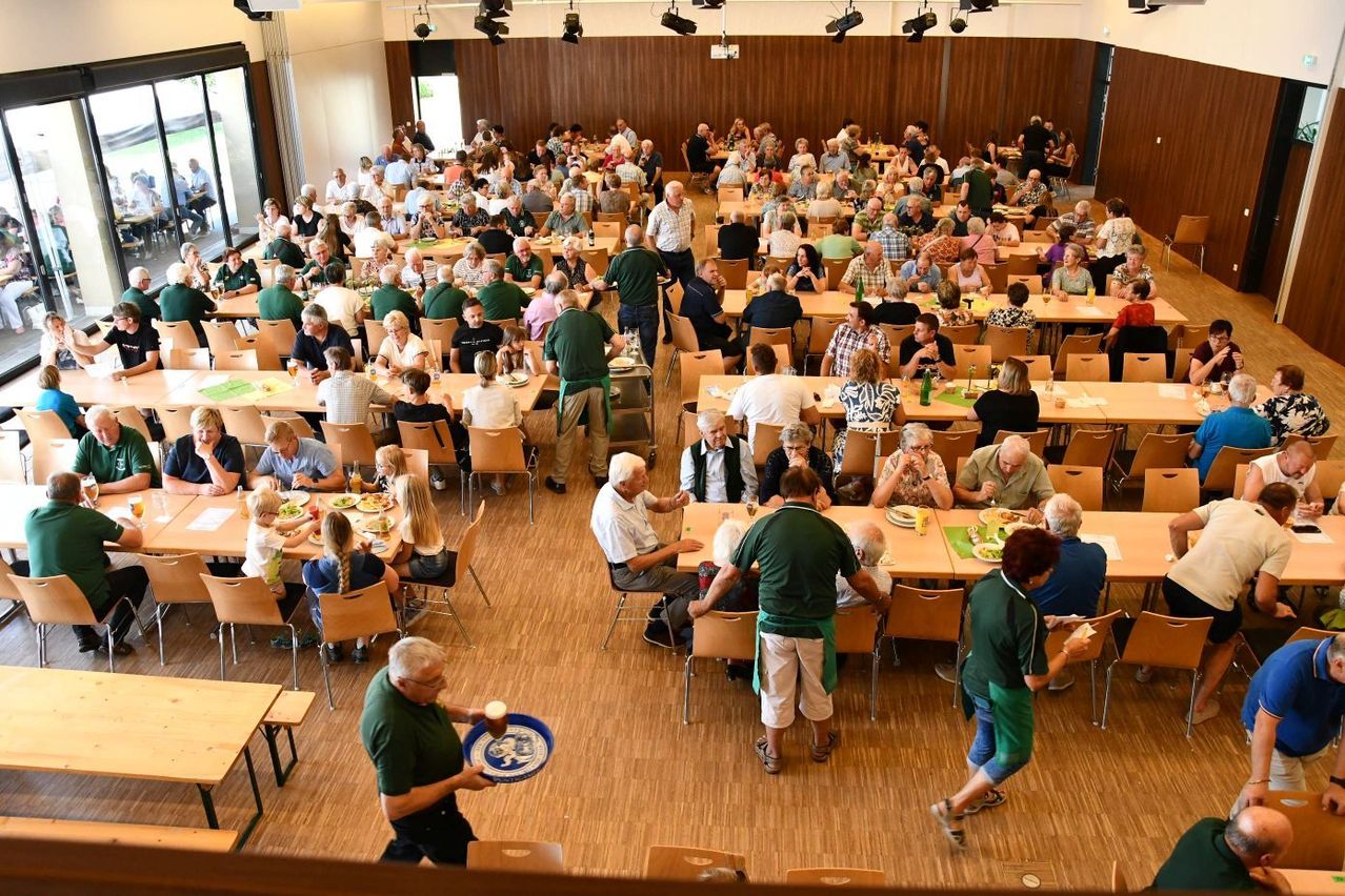 A large dining hall filled with elderly people. They are seated at long tables, some are standing. A man is holding a tray with a cup.