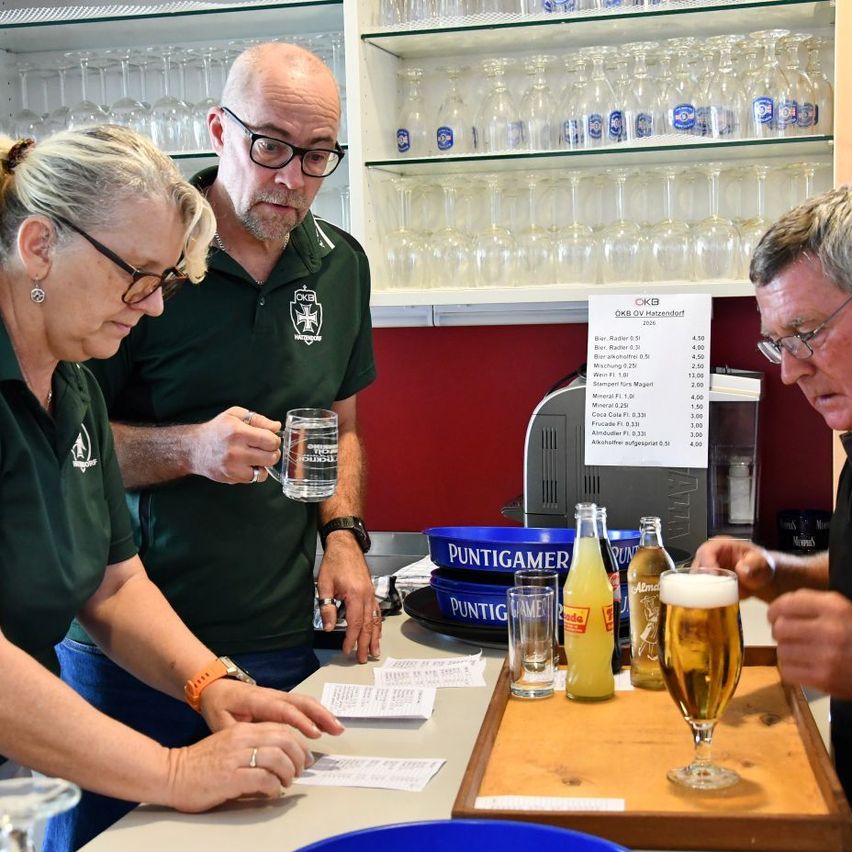 Three people in green polo shirts are at a bar counter. They have glasses and bottles of beer on the counter. A price list is on the wall.