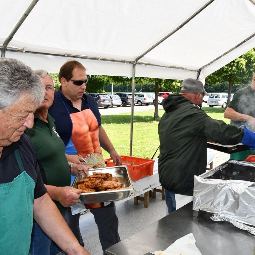Several people are preparing food in an outdoor tent, under a white canopy. A man with a blue apron is holding a tray of food. Nearby, a man is grilling food. There are cars parked in the distance.