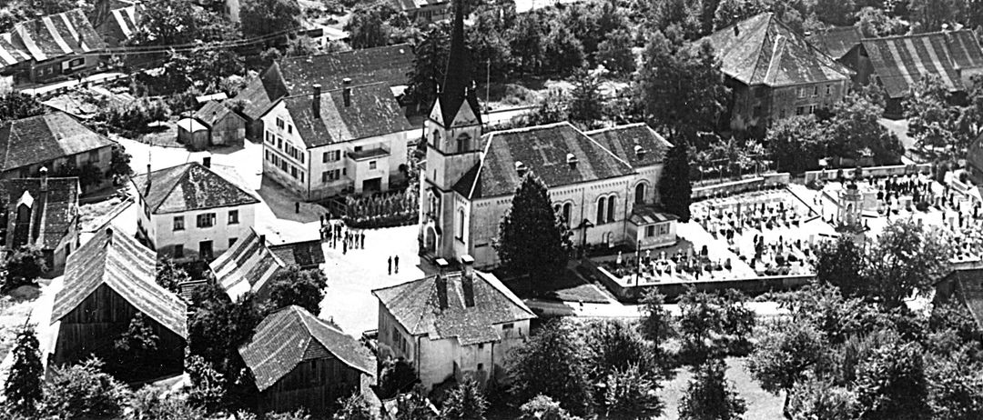 Ein altes Luftbild eines Dorfes mit einer Kirche, einer Brücke über einem Fluss und einer Straße, die dorthin führt. Bäume und Häuser umgeben das Gebiet.