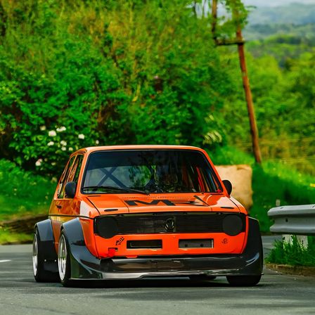 Ein orangefarbenes Rennauto mit schwarzen Akzenten fährt auf einer Straße mit Grünflächen und einem Schutzgeländer.